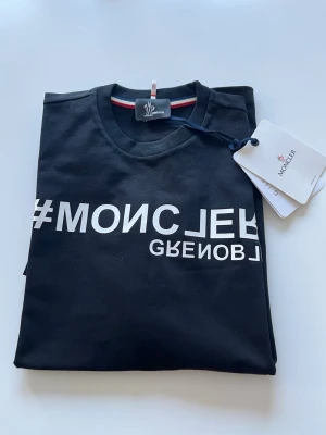 Moncler t-shirt  - Snygg svart t-shirt från Moncler Grenoble med vit textprint framtill. Klassisk rund halsringning och diskret Moncler-logga på etiketten. T-shirten har en stilren design och är tillverkad i mjuk bomull för en bekväm känsla.