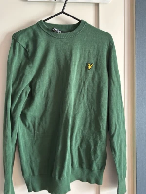 Grön tröja från Lyle & Scott - Grön långärmad tröja från Lyle & Scott med rund halsringning och klassisk gul logga på bröstet. Tröjan är tunnstickad och har ribbade muddar vid ärmslut och nederkant. Perfekt för lager-på-lager och enkel att matcha.