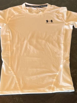 Vit långärmad kompressionströja från Under Armour - Helt ny och oanvänd. Snygg vit långärmad träningströja från Under Armour med svart logga på bröstet. Tillverkad i ett stretchigt och fukttransporterande material som passar perfekt för intensiva träningspass. Tight passform och rund halsringning. 