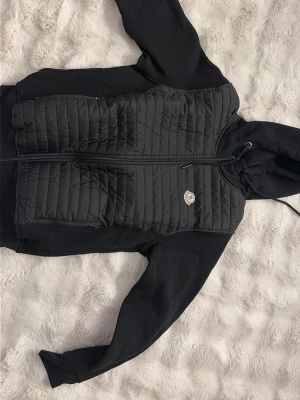 Svart quiltad jacka från Moncler - Svart jacka från Moncler med quiltad framsida, huva med snörning och dragkedja. Jackan har långa ärmar i mjukt material och en liten logotyp på bröstet. Perfekt för kyliga dagar och snygg till streetwear. Fick den i present men kommer aldrig till användning då jag har två andra