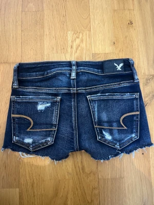 American Eagle shorts  - Snygga mörkblå jeansshorts från American Eagle med slitna detaljer och råa kanter. Klassisk femficksmodell med låg midja och coola kontrastsömmar på bakfickorna. Perfekta för en avslappnad stil under varma dagar. Skulle säga att dom är Stl 24