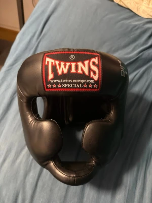 Svart huvudskydd från Twins Special - Säljer ett svart huvudskydd från Twins Special, perfekt för boxning eller kampsport. Tillverkat i slitstarkt konstläder med vaddering runt hela huvudet och öppning för ansiktet. Justerbar passform med kardborreband. Klassisk design med röd och vit logga framtill.