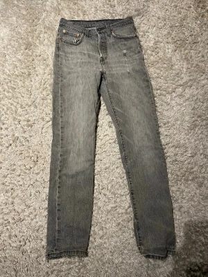 Grå jeans från Levi's - Säljer ett par grå jeans från Levi's med klassisk femficksdesign och raka ben. Jeansen har en snygg tvättad look och Levi's ikoniska läderpatch bak i midjan. Perfekta för dig som gillar en stilren och avslappnad vibe. Har en liten slitning. Skulle säga att storleken är runt L30 W28.