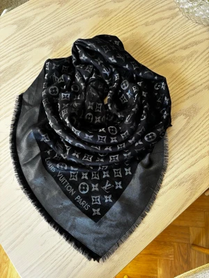 Svart Louis Vuitton sjal med monogram - Snygg svart sjal från Louis Vuitton med ikoniskt monogrammönster i grått och svart. Tillverkad i en lyxig mix av silke, metall och ull. Sjalen har fransade kanter och känns riktigt exklusiv. Köpt i en vintage butik så inget kvitto. Mycket fint skick. 