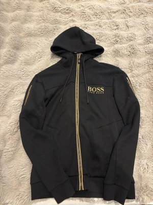 Svart hoodie från Hugo Boss - Snygg svart hoodie från Hugo Boss med guldfärgade detaljer och dragkedja. Märket syns tydligt på bröstet och ärmen. Hoodien har huva med dragsko och ribbade muddar. Perfekt för en stilren och modern look.