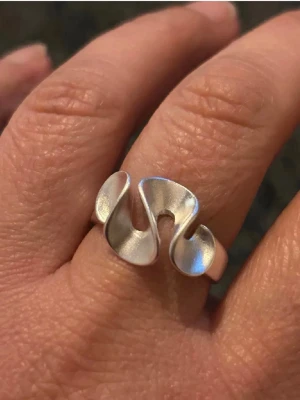 Silvrig vågig ring i modern design - Unik ring i silver metall med vågig, böljande form som ger ett coolt och trendigt intryck. Ringen är öppen baktill vilket gör den justerbar och passar flera storlekar. Perfekt för dig som gillar statement-smycken och modern stil.
