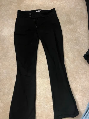 Svarta bootcut byxor med knappar - Säljer ett par svarta bootcut byxor med två knappar i midjan och lätt utsvängda ben. Byxorna har en klassisk look och passar perfekt till många olika stilar. Materialet känns mjukt och stretchigt.