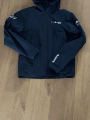 Mörkblå Spray Gore Tex vindjacka - Snygg mörkblå vindjacka från Sail Racing med huva och dragkedja framtill. Tillverkad i slitstarkt Gore Tex-material som står emot både vind och regn. Jackan har logotyper på bröstet, ärmen och handleden samt två praktiska sidofickor med dragkedja. Original pris 3500 kr.