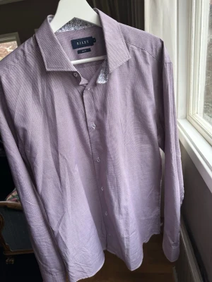 Ljuslila skjorta från Riley - Snygg ljuslila skjorta från Riley med diskret rutigt mönster och vita knappar. Skjortan har klassisk krage och lång ärm, samt ett blommigt tyg på insidan av kragen för en extra detalj. Perfekt för dig som gillar stilrena plagg med en twist.