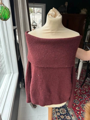 Vinröd off shoulder stickad tröja - Mysig vinröd stickad tröja med bred off shoulder-krage. Tröjan har en avslappnad passform och ribbstickad kant nedtill. Perfekt för dig som vill ha en trendig och bekväm look till hösten eller vintern.