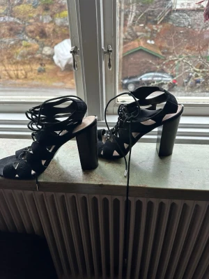 Svarta snörade pumps med klack - Säljer ett par svarta pumps med höga blockklackar och snygga snörningsdetaljer framtill. Skorna har öppna partier på sidorna och är tillverkade i mockaimitation. Perfekta för dig som vill ha en edgy och trendig look.