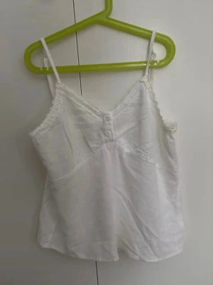 Vit spetskantad linnetopp - Söt vit linnetopp med tunna axelband och spetskant längs urringningen. Toppen har små  knappar framtill och är i linnetyg. Helt ny och aldrig använd