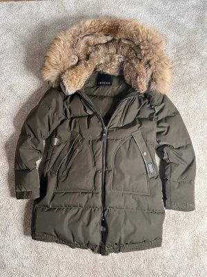 Mörkgrön pufferjacka från Cedrico - Snygg mörkgrön pufferjacka från Cedrico med stor fluffig päls på huvan och perfekt för de kalla dagarna nu för vintern. Jackan har dragkedja framtill, flera fickor med dragkedja och snörning nedtill och har en riktigt schysst oversized look. Prutat och klart. Skriv efter minsta lilla fundering!!