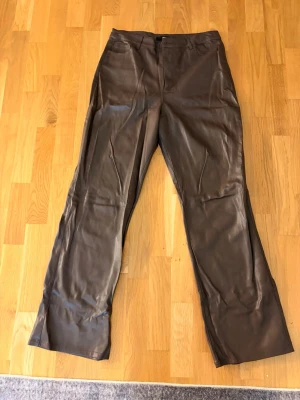 Bruna fuskskinnbyxor från BikBok - Snygga bruna byxor i fuskskinn från BikBok. Byxorna har raka ben, hög midja och stängs med knapp och dragkedja. Knappen lite lös.Perfekta för dig som vill ha en trendig och edgy look. Materialet ger en cool läderkänsla och passar till många olika stilar.