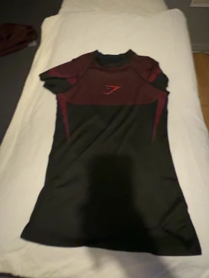 Gymshark Onyx - Snygg svart och vinröd träningströja från Gymshark med korta ärmar och sportig passform. Tillverkad i ett lätt och stretchigt funktionsmaterial som andas, perfekt för gymmet eller löpning. Gymshark-logga på bröstet.