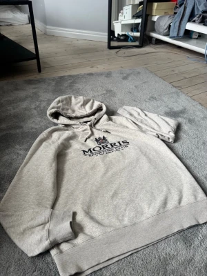 Grå hoodie från Morris Sporting Club - Snygg ljusgrå hoodie från Morris Sporting Club med broderad logga och text på bröstet. Klassisk huva med dragsko och ribbade muddar vid ärmslut och nederkant. Perfekt för en avslappnad och stilren look.