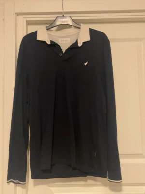 Mörkblå långärmad henley från pier one - Mörkblå långärmad henley från pier one med kontrasterande ljus krage och knappslå. Liten vit logga på bröstet och diskreta vita detaljer vid ärmsluten. Perfekt för en avslappnad men stilren look.