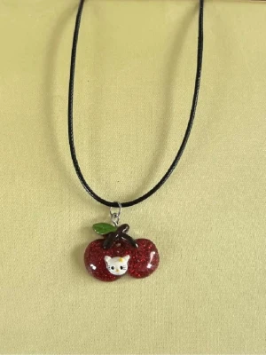Halsband med körsbär och katt - Sött halsband med svart snöre och ett hänge i form av två glittriga röda körsbär med ett grönt blad och en liten vit katt i mitten. Perfekt för dig som gillar gulliga accessoarer och vill sticka ut lite extra.