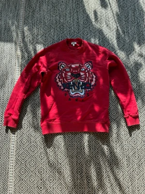 Röd sweatshirt med tiger från Kenzo - Cool röd sweatshirt från Kenzo med det ikoniska tigermotivet broderat på bröstet och texten 'Kenzo Paris' under. Tröjan har rund hals, ribbade muddar och är gjord i mjuk bomull. Perfekt för dig som vill sticka ut med en statement-piece.