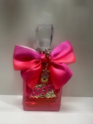Juicy Couture Viva La Juicy parfym Neon - Juicy Couture Viva La Juicy parfym i en fyrkantig rosa glasflaska med en stor dekorativ rosett i rosa och cerise satin. Flaskan har ett genomskinligt, kristalliknande lock och ett guldfärgat hänge med logga och texten 'Juicy'. Perfekt för dig som gillar färgstark design, som helt ny.