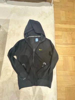 Svart Nike hoodie med gul logga - Cool svart hoodie från Nike med gul swoosh på bröstet. Tröjan har dragkedja, huva och två fickor framtill. Materialet är mjukt och bekvämt, perfekt för chill eller träning. Snygg design med diskreta detaljer och sportig känsla.
