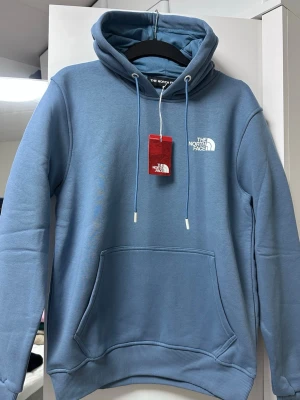 Blå hoodie från The North Face - Snygg blå hoodie från The North Face med vit logga på bröstet. Klassisk känguruficka framtill och justerbar huva med dragsko. Tillverkad i mjukt material som känns skönt mot huden. Perfekt för en avslappnad och trendig look.