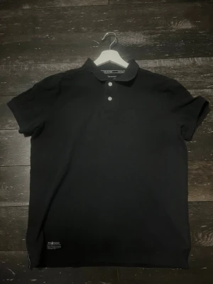 Mörkblå pikétröja från Stone Island - Klassisk mörkblå pikétröja från sail racing med korta ärmar och två knappar framtill. Tröjan har en liten logotyp på ärmen och är tillverkad i mjuk bomull för en bekväm känsla. Perfekt till jeans eller shorts för en clean look. Hör av er vid frågor!