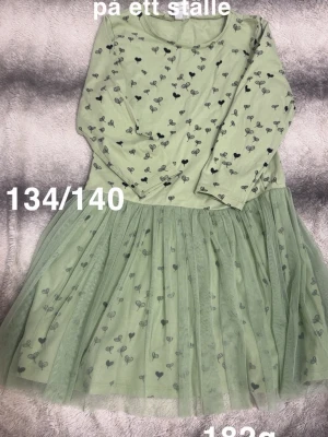 Grön långärmad klänning med hjärtan - Söt grön klänning med långärmad överdel och tyllkjol. Klänningen har små svarta hjärtan som mönster över hela plagget och en mjuk, flowy kjol. Perfekt för dig som gillar gulliga detaljer och bekväma klänningar.🌸 Har blivit en reva i tyllen. Storlek 134/140 