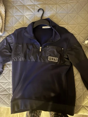 Mörkblå half-zip hoodie från Valentino - Snygg mörkblå hoodie från Valentino med half-zip och VLTN-logga på bröstet. Materialmix med glansig panel över bröstet och mjuk bomull i resten av tröjan. Hög krage och ribbade muddar för extra stilpoäng. Perfekt för dig som gillar exklusiva streetwear-plagg.