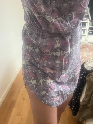 Lila mönstrad kortklänning med bälte - Kortklänning från Didriksons i lila och rosa toner med abstrakt mönster. Klänningen har korta ärmar, knappar framtill och ett matchande bälte i midjan. Perfekt för sommarens alla tillfällen och tillverkad i mjuk bomull.