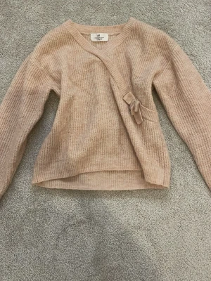 Beige omlottstickad tröja från H&M - Storlek 122/128 men tycker själv den passar som 140/146, jag själv brukar ha 146/152 eller 158 och denna passar perfekt men den är bara lite kort i armarna! Tröjan ser beige ut på bilden men egentligen är den mer rosa, stickad omlott-tröja från H&M med ribbad struktur och knytband i sidan. Tröjan har V-ringning och långa ärmar, perfekt för lager på lager och en chill look. Materialet är mjukt och skönt, passar till det mesta i garderoben.