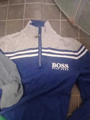 Blå och grå Hugo Boss tröja - Snygg blå och grå tröja från Hugo Boss med vit logga på bröstet och vita ränder över bröstet. Tröjan har halv dragkedja och hög krage, perfekt för en sportig och avslappnad stil.