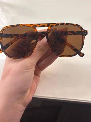Grish solglasögon  - Snygga solglasögon med leopardmönstrad båge i brunt och svart. Modellen har stora, fyrkantiga glas och bruna linser som ger en cool retrovibe. Perfekt accessoar för att lyfta din stil i solen.