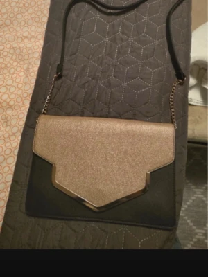 Svart och guld kuvertväska - Kuvertväska i svart och guld med geometrisk form på locket och metallkant. Väskan har en axelrem i svart och kedjedetaljer i guld. Materialet ser ut att vara syntet med glittrigt lock. Perfekt för att lyfta din outfit , den är ny.