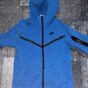 Blå Nike techfleece  - Blå techfleece från Nike med hel dragkedja framtill och svart Nike-logga på bröstet. Tröjan har huva, svarta detaljer längs bröstet och på ärmen samt praktiska fickor. Perfekt för en sportig och avslappnad stil.