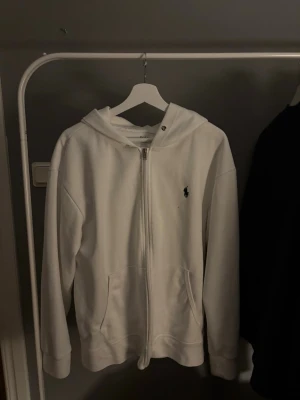Vit hoodie från Polo Ralph Lauren - Snygg vit hoodie från Polo Ralph Lauren med dragkedja framtill och klassisk huva. Har två fickor och den ikoniska lilla loggan broderad på bröstet. Perfekt för en clean och stilren look. Tillverkad i mjukt bomullsmaterial.