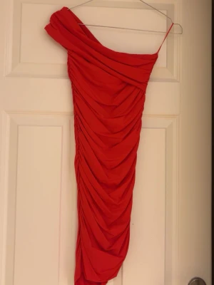 Röd oneshoulder klänning med drapering - Säljer en snygg röd oneshoulder klänning med draperad design och smal passform. Klänningen har en axelbandslös sida och ett smalt axelband på andra sidan. Perfekt för dig som vill sticka ut med en trendig look.