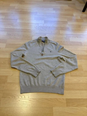 Beige halvzip-tröja från Morris - Snygg beige tröja från Morris med halvzip och liten broderad logga på bröstet. Tröjan har ribbade muddar vid ärmslut och nederkant samt en klassisk ståkrage. Perfekt för en stilren och avslappnad look.