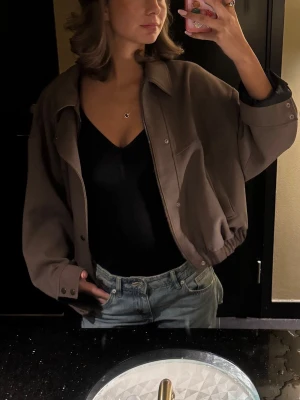 Brun bomberjacka med knappar - Snygg brun bomberjacka med avslappnad passform och knappar framtill. Jackan har långa ärmar, klassisk krage och resår i midjan för en cool siluett. Perfekt att slänga över en t-shirt eller hoodie.