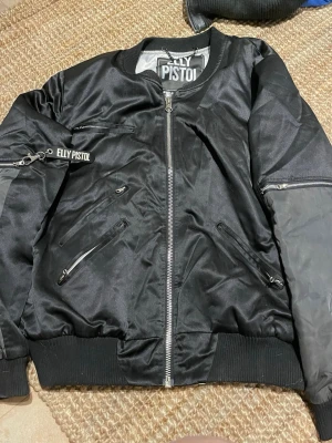 Svart bomberjacka från Elly Pistol - Snygg svart bomberjacka från Elly Pistol med silvriga dragkedjor och ribbade muddar. Jackan har flera fickor med dragkedja och en cool detalj med Elly Pistol-nyckelband på ärmen. Perfekt för en streetstyle-look.