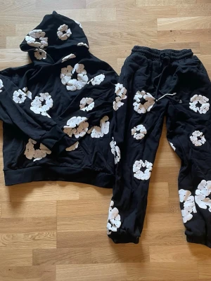 Denim tears hoodie och byxor - Säljer ett svart loungewear-set med vita blommor. Setet består av en hoodie med huva och matchande byxor med resår i midjan och benslut. Perfekt för chill dagar hemma eller när du vill vara bekväm men ändå snygg.