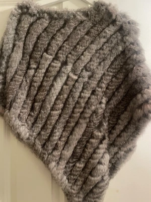 Grå pälsväst - Supermjuk och fluffig grå sjal i fuskpäls med diagonala ränder. Sjalen har en trekantig form och ger en lyxig känsla till din outfit. Perfekt att svepa om sig när det är kyligt ute och vill ha något extra snyggt.