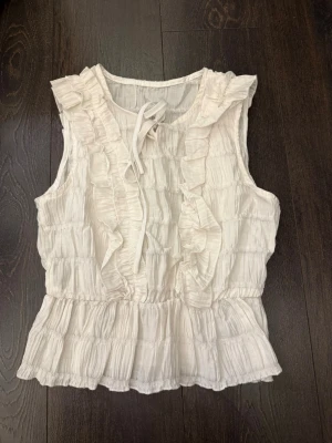 Vit ärmlös blus med volanger - Söt vit blus i luftig och lätt material med volangdetaljer framtill och knytband vid halsen. Blusen är ärmlös och har en rynkad struktur som ger en romantisk känsla. Perfekt att matcha med jeans eller kjol för en trendig look.