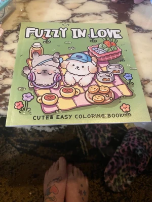 Fuzzy In Love målarbok - En söt och lätt målarbok med titeln 'Fuzzy In Love'. Omslaget har gulliga djurfigurer på picknick, tecknade i pastellfärger. Boken har mjukt omslag och innehåller flera illustrationer att färglägga, perfekt för dig som gillar kawaii och kreativa aktiviteter.
