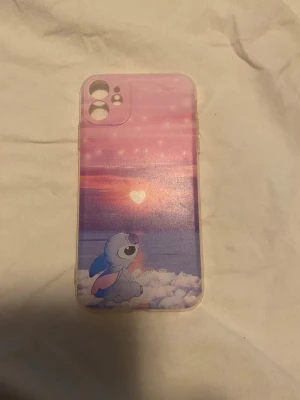 Rosa mobilskal med Stitch och solnedgång - Säljer ett mobilskal med motiv av Stitch som sitter på ett moln och tittar på en rosa-lila solnedgång med ett hjärta i mitten. Skalets färger går i rosa, lila och blått och det är gjort i plast. Perfekt för dig som gillar söta och färgglada accessoarer till mobilen. Passar IPhone 11