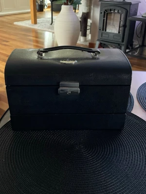 Svart smyckeskrin i läderimitation - Stilrent svart smyckeskrin i läderimitation med struktur, silverfärgat lås och handtag. Perfekt för att organisera och förvara dina smycken. Klassisk design med rundad topp och metallbeslag för en lyxig känsla.