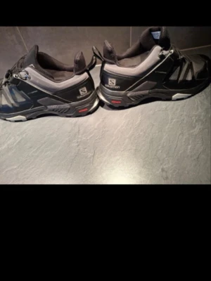 Svarta Salomon sneakers med grå detaljer - Säljer ett par svarta Salomon sneakers med grå inslag och robust sula med grovt mönster. Skorna har låg profil och snabbsnörning, perfekt för dig som gillar en sportig och funktionell stil. Materialet är syntet och mesh för bra ventilation. Storlek 44 men passar 43