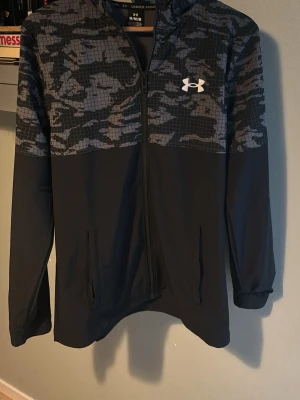 Svart vindjacka från Under Armour - Snygg svart vindjacka från Under Armour med grått camomönster upptill och vitt logotryck på bröstet. Jackan har huva, dragkedja framtill och två fickor. Tillverkad i lätt och smidigt material, perfekt för blåsiga dagar.