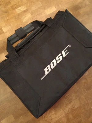 Svart väska från Bose med handtag - Säljer en svart väska från Bose med tydlig logga på framsidan. Väskan har två robusta handtag med vaddering för extra komfort. Tillverkad i slitstarkt tygmaterial och har en rektangulär form, perfekt för att bära tillbehör eller utrustning. Väskan har en kardborre stängning.