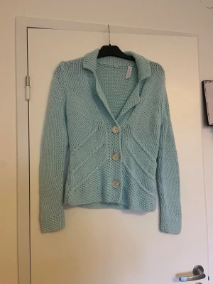 Ljusblå stickad kofta med stora knappar - Snygg ljusblå stickad kofta med tre stora knappar framtill och bred krage. Koftan har ett unikt mönster med diagonala stickningar och långa ärmar. Perfekt att slänga på sig när det är lite kyligt ute.